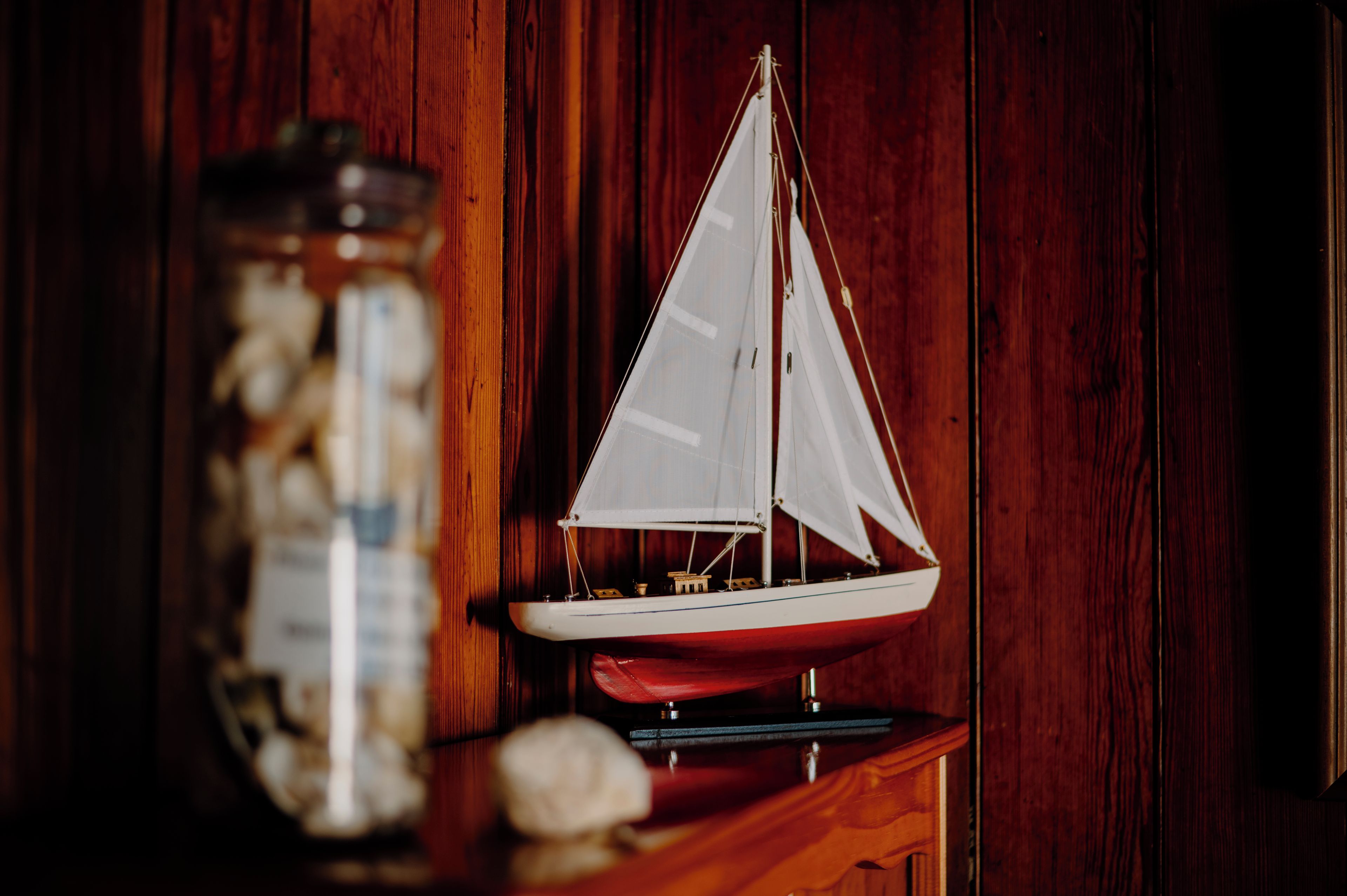 Een modelzeilboot op een houten ondergrond, met een pot met spullen op de achtergrond.