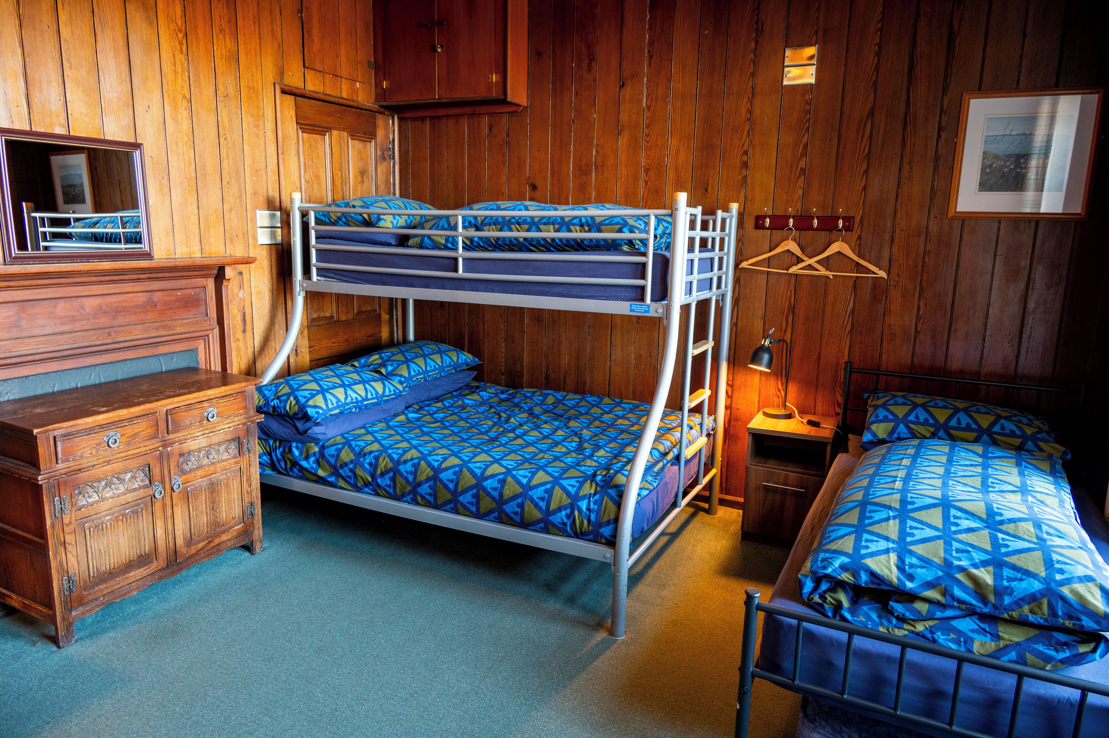 A bunk bed with patterned bedding, a wooden dresser, a bedside table with a lamp, and a framed picture on the wall.