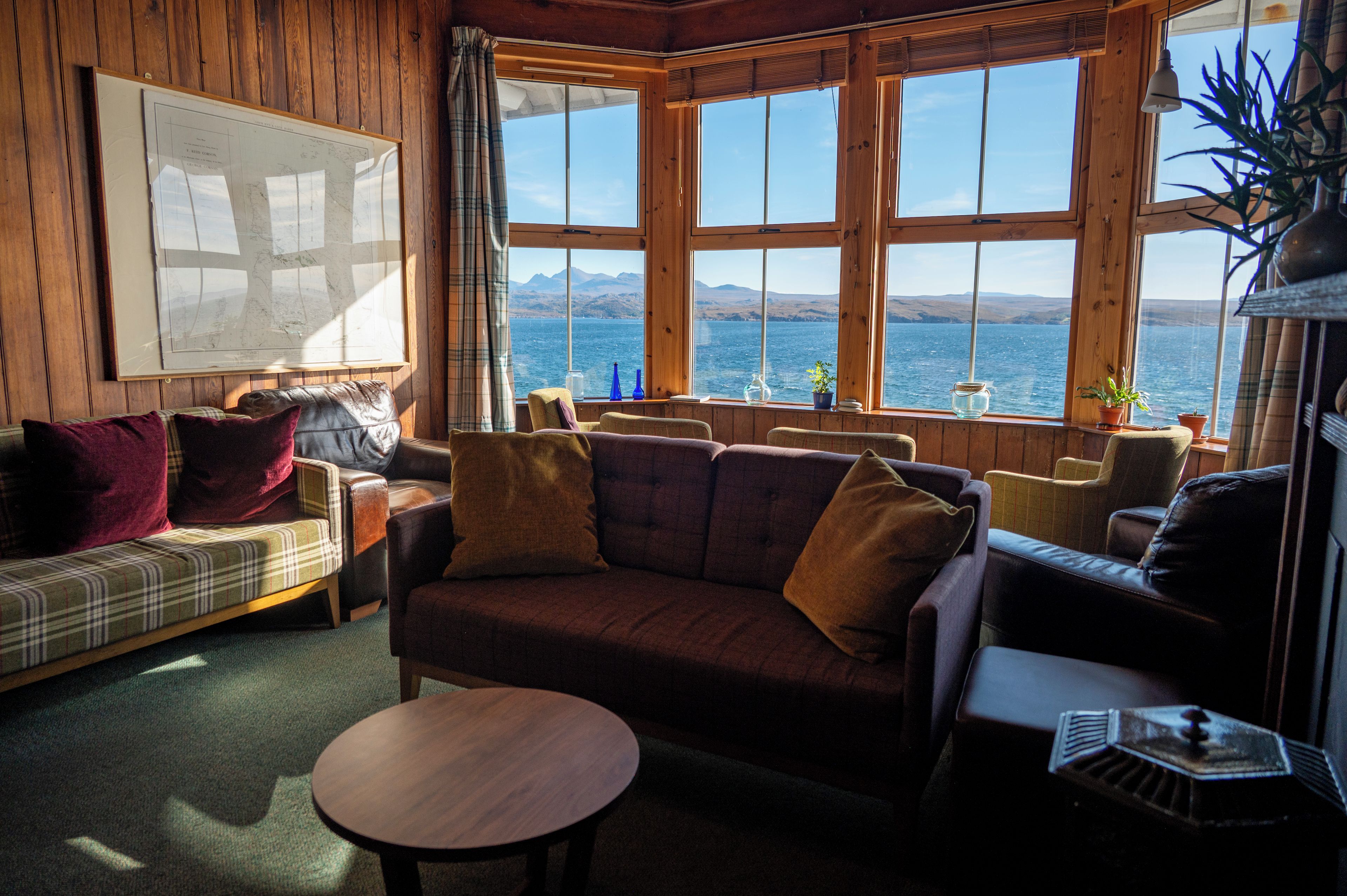 A cozy cabin interior with a large window offering a view of the sea and hills, a map on the wall, and a mix of leather and fabric furniture.
