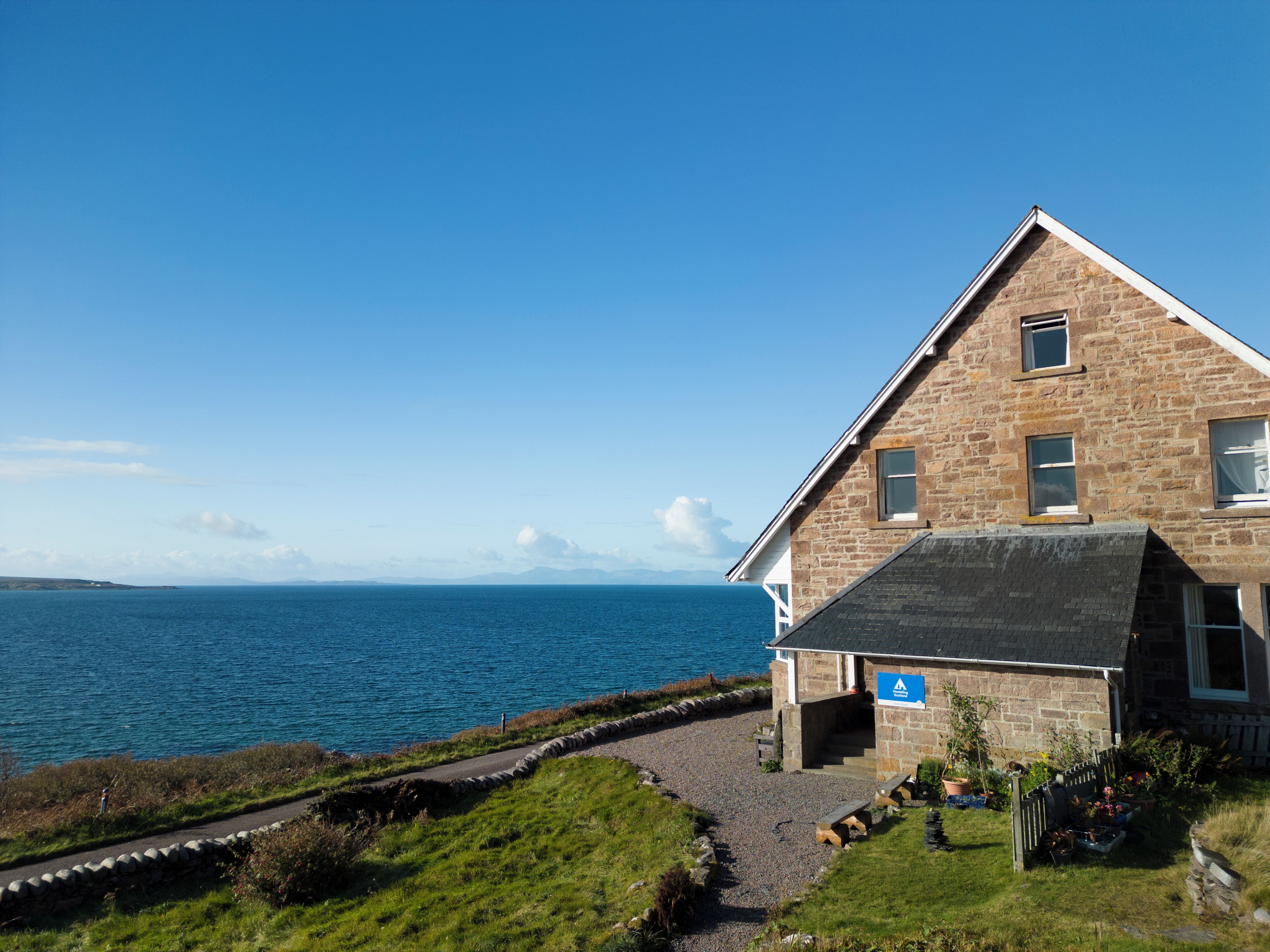 A stone building with a sloped roof, a gravel driveway, and a clear view of the sea.