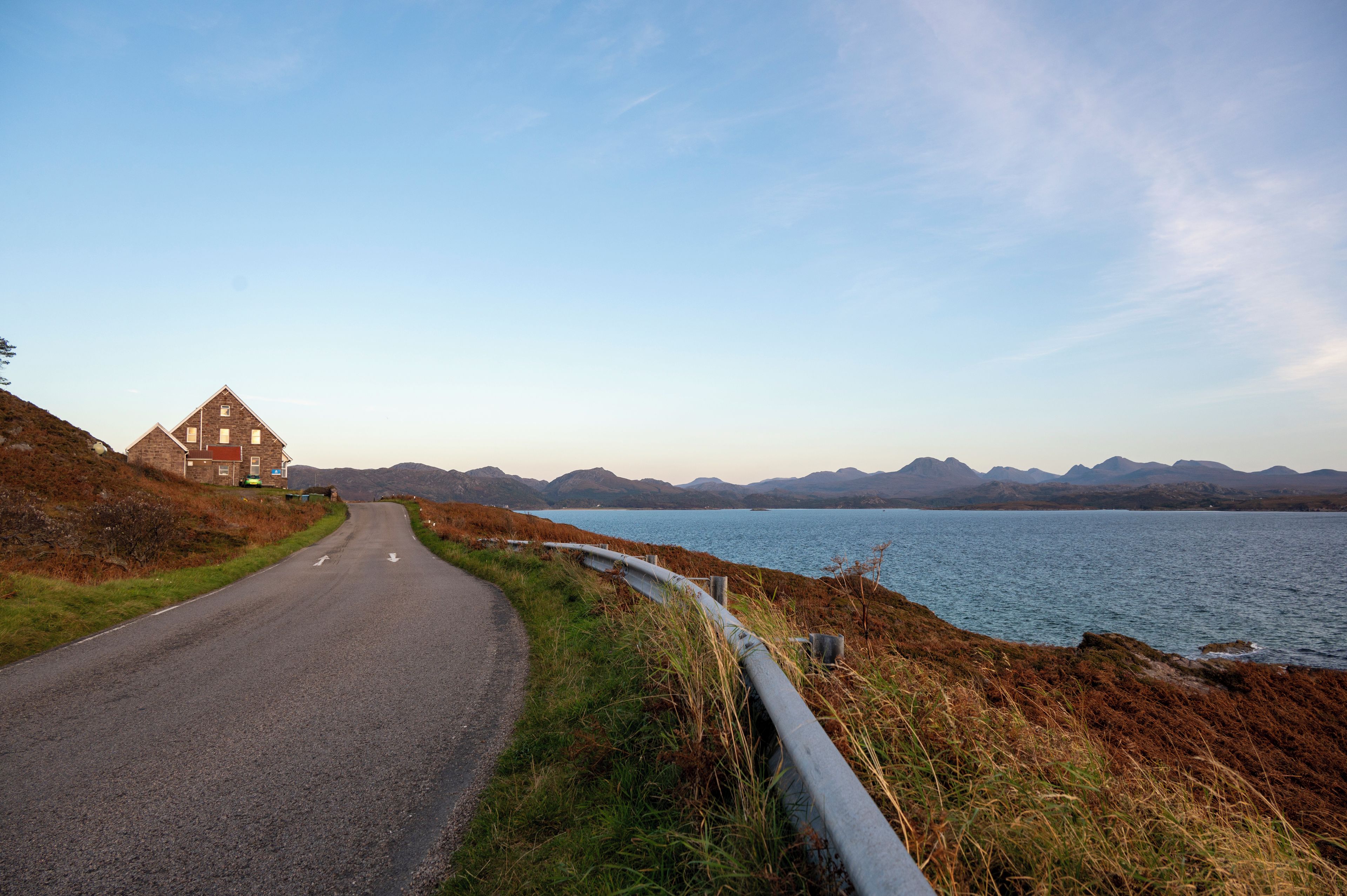 A road leading to a stone house with a view of a lake and mountains in the distance.