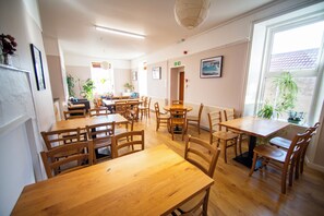 A dining area with multiple wooden tables and chairs, a ceiling light fixture, and a window with a view of the outside. - Gairloch Sands Youth Hostel (Gairloch)