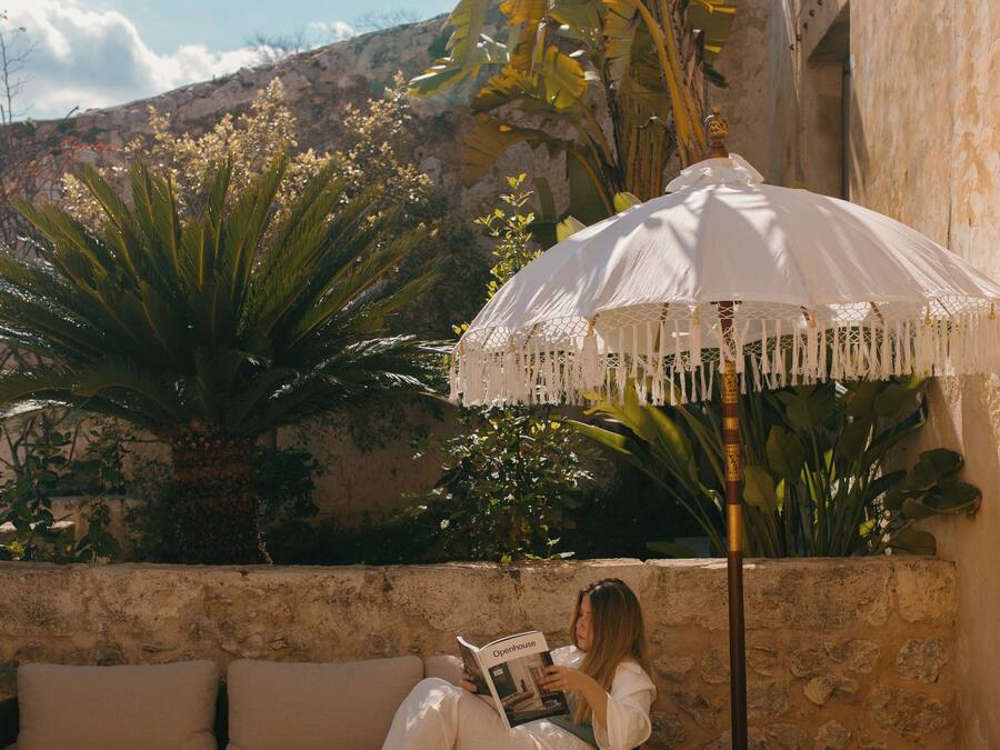 A woman reading under a white umbrella in a courtyard with a stone wall, a sofa, and a wooden table.