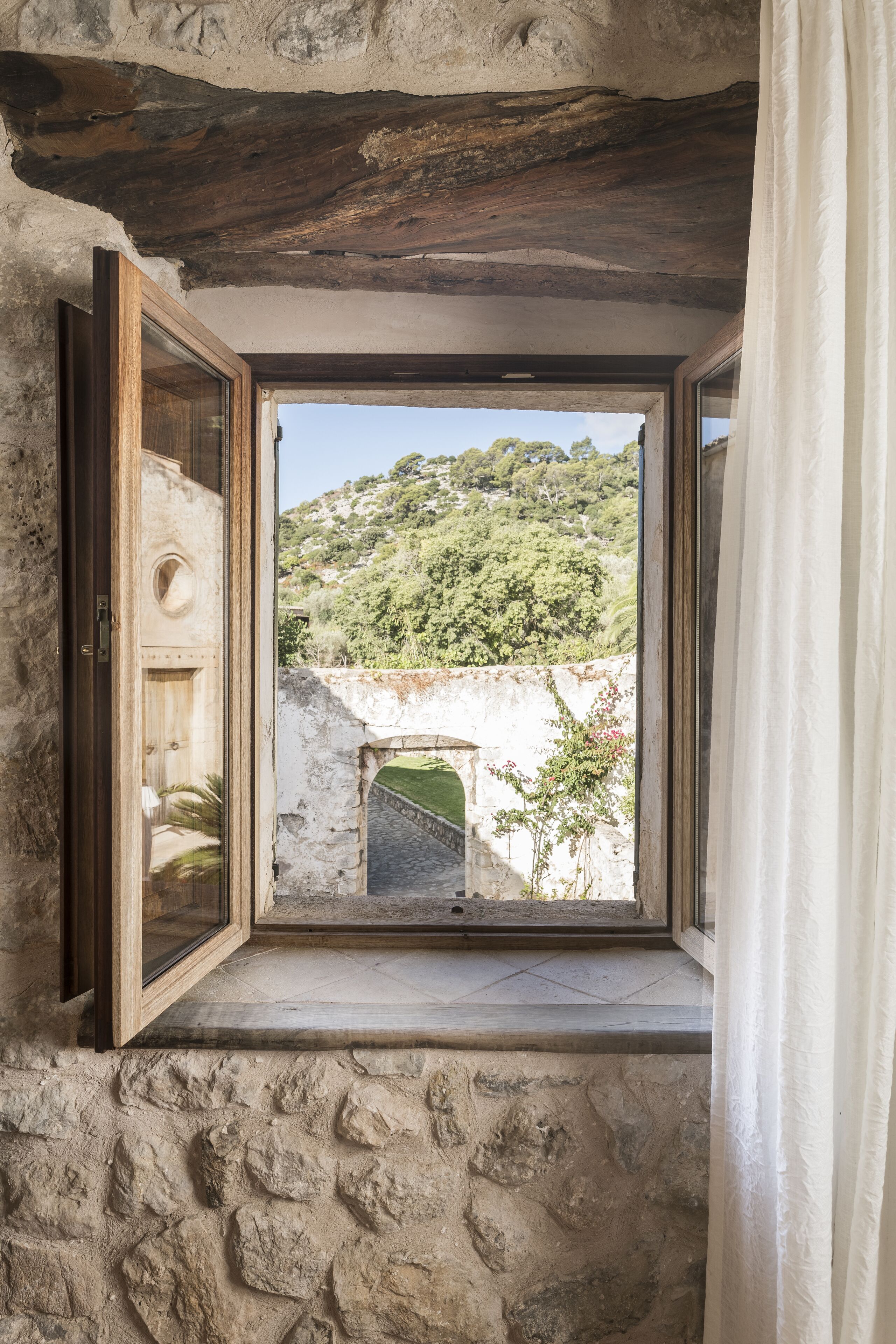 A stone-walled room with a large window overlooking a lush hillside and an ancient stone bridge.