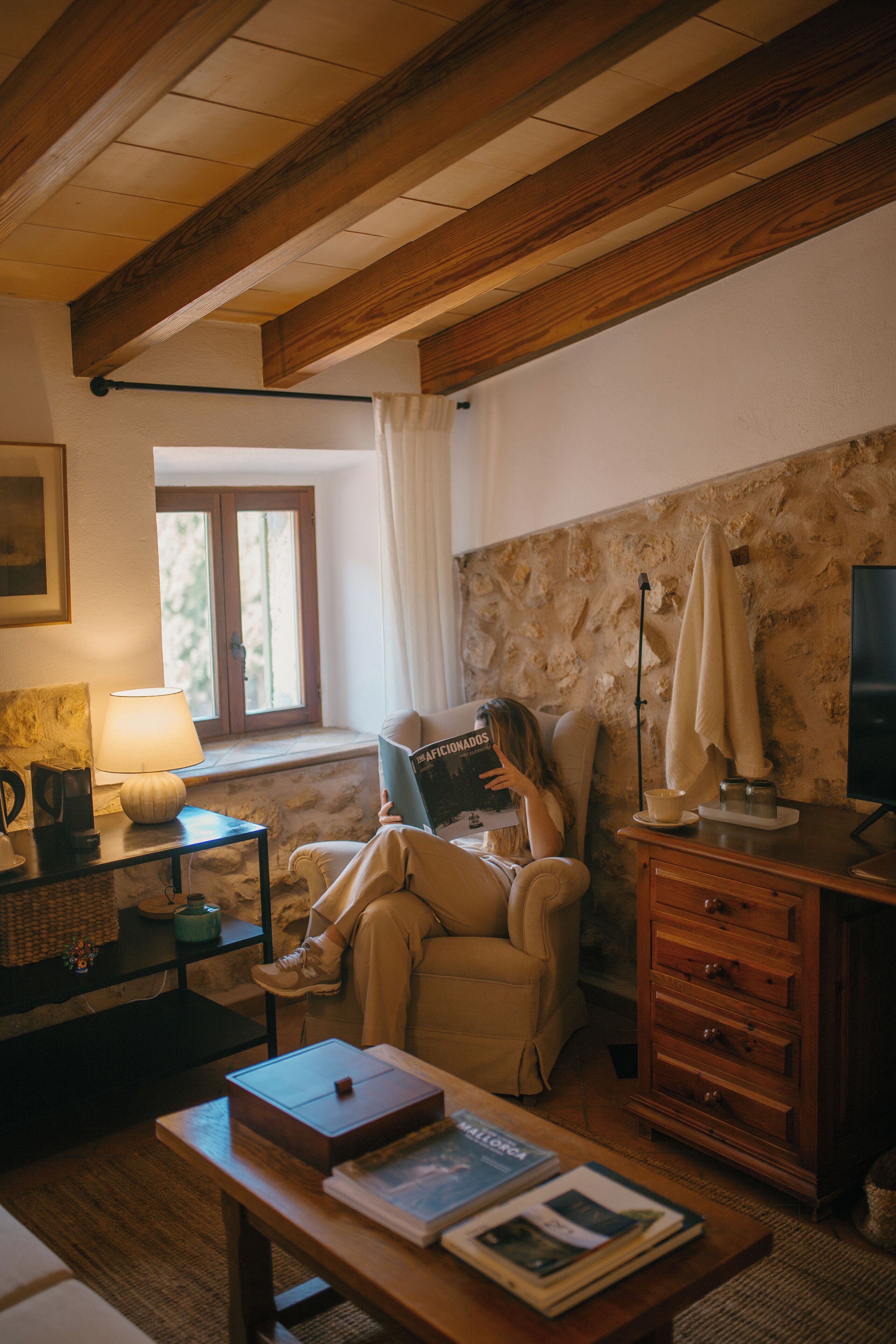 Una habitación acogedora con techo de madera, una ventana con cortinas, una lámpara, un sofá, un televisor y una estantería.
