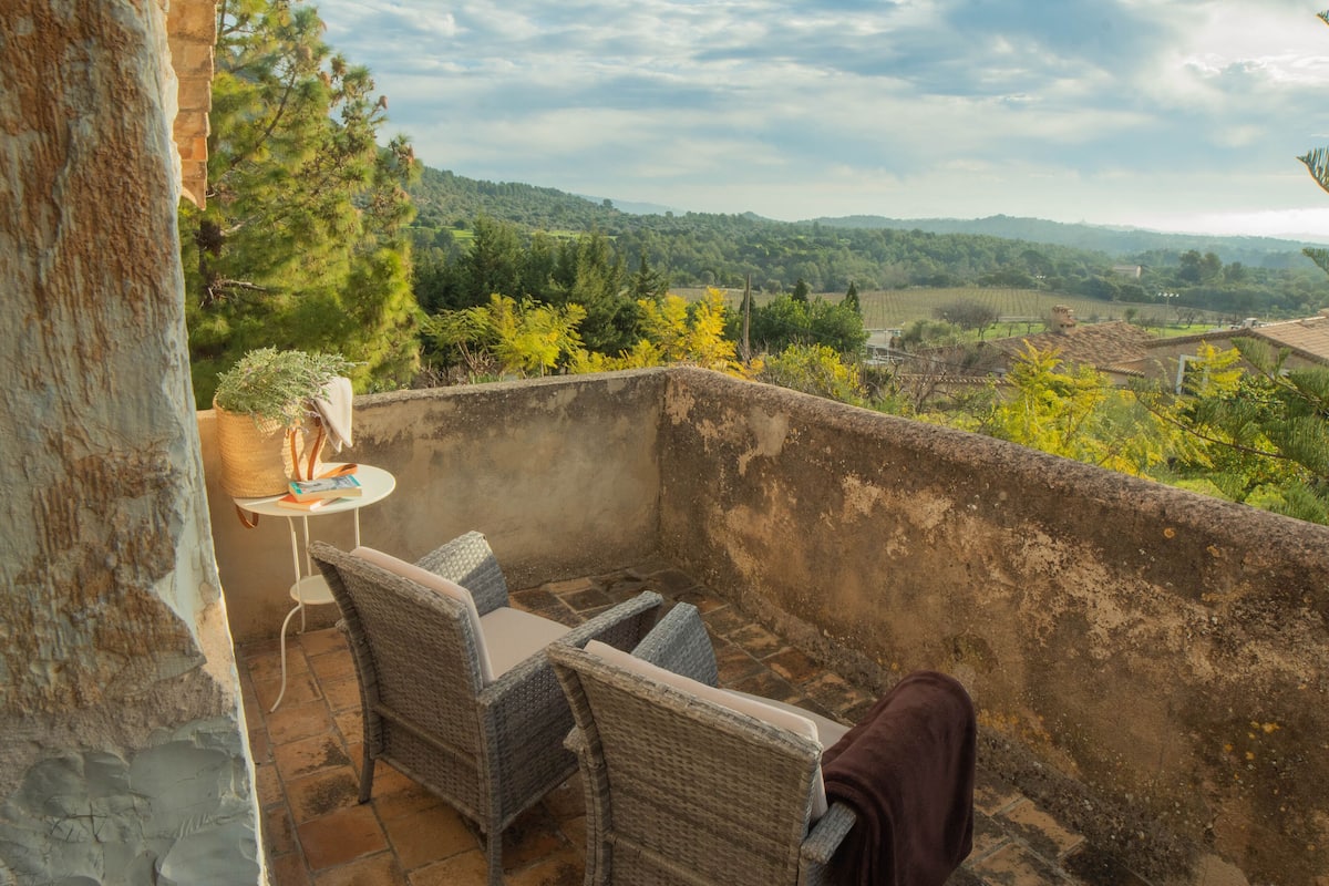 A balcony with wicker chairs and a small table, overlooking a scenic landscape with trees and hills.