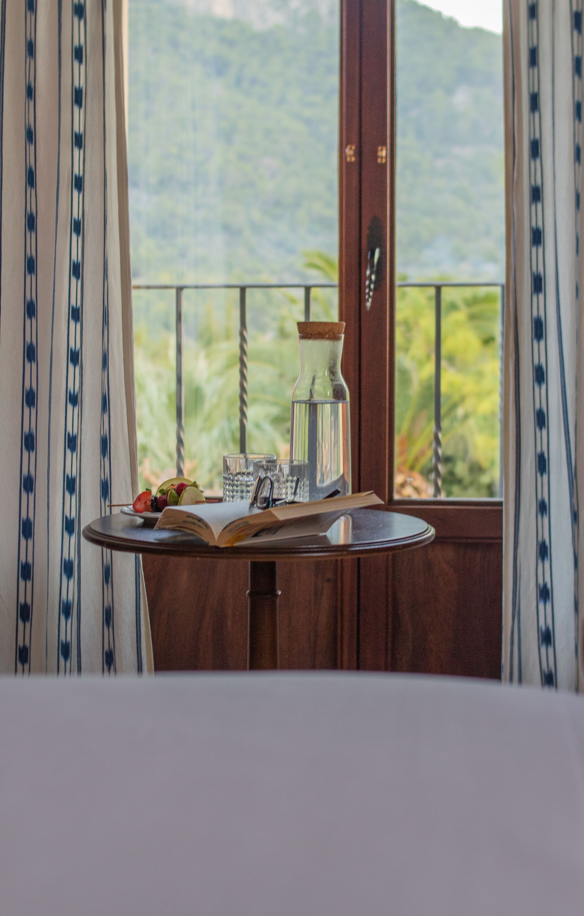 A table with a book, a glass, and a bottle, set against a window with a view of greenery and mountains.