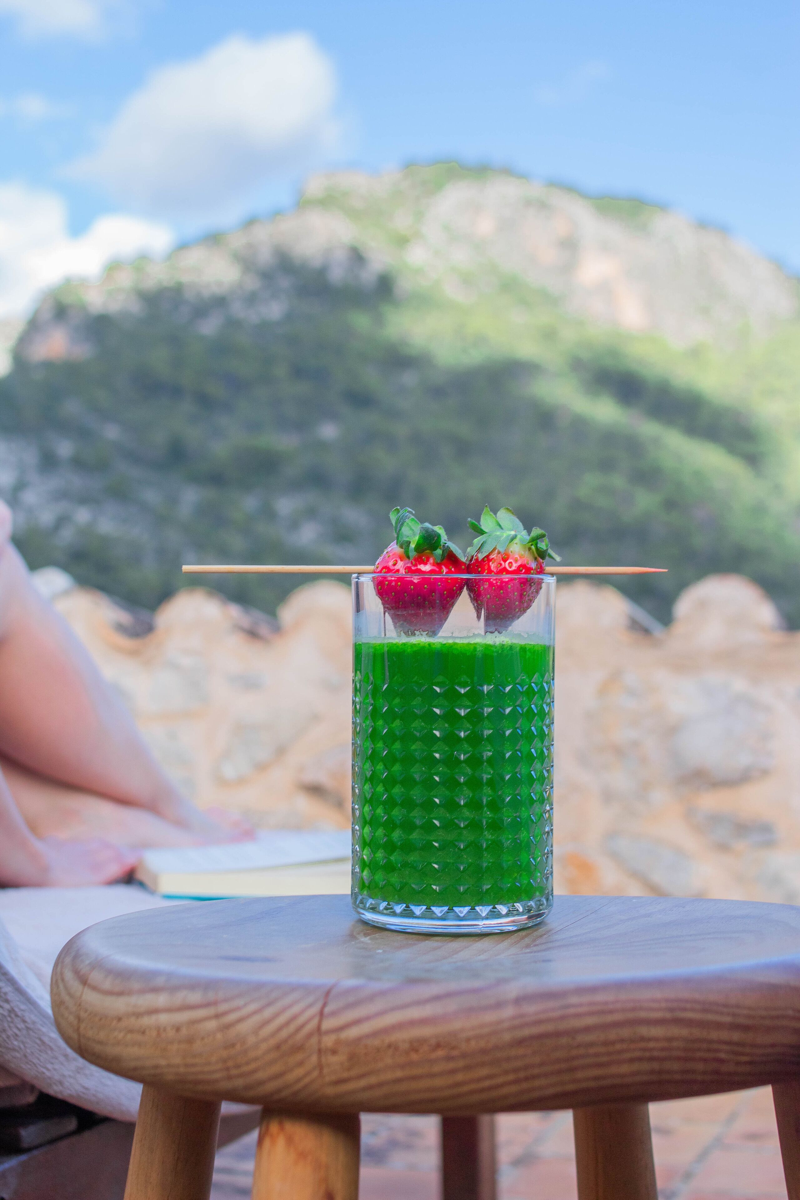 Un verre d’une boisson verte, garni de fraises, est posé sur une table en bois, avec une vue sur les montagnes en arrière-plan.