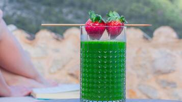 Un verre d’une boisson verte, garni de fraises, est posé sur une table en bois, avec une vue sur les montagnes en arrière-plan.