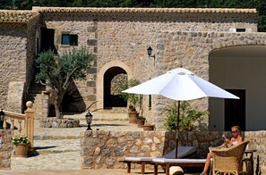 A stone building with a tiled roof, a staircase leading to an entrance, and outdoor seating with a white umbrella. -  Finca Can Beneït (Selva)
