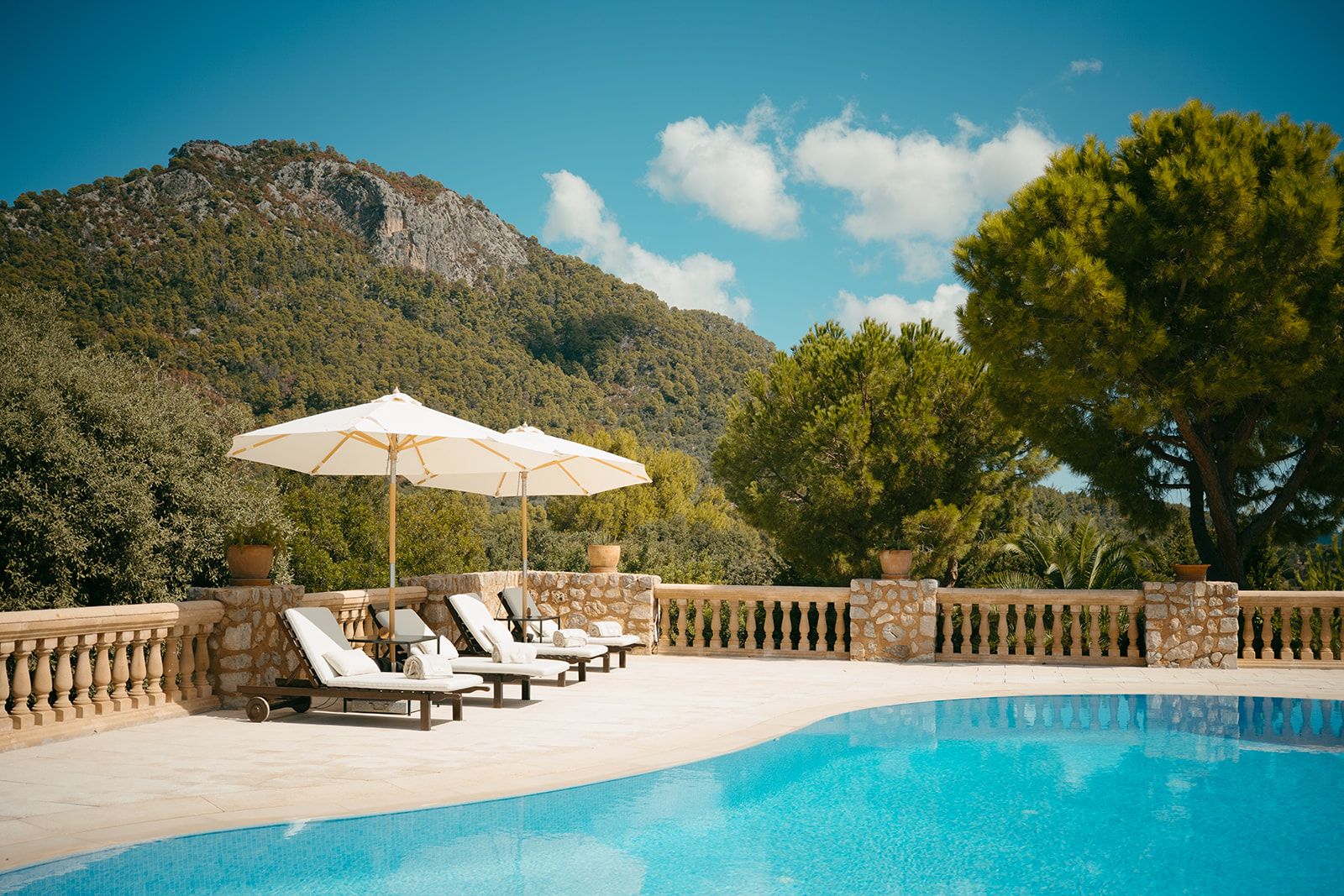 Un área de piscina con sillones y sombrillas, rodeada de muros de piedra y un paisaje montañoso.