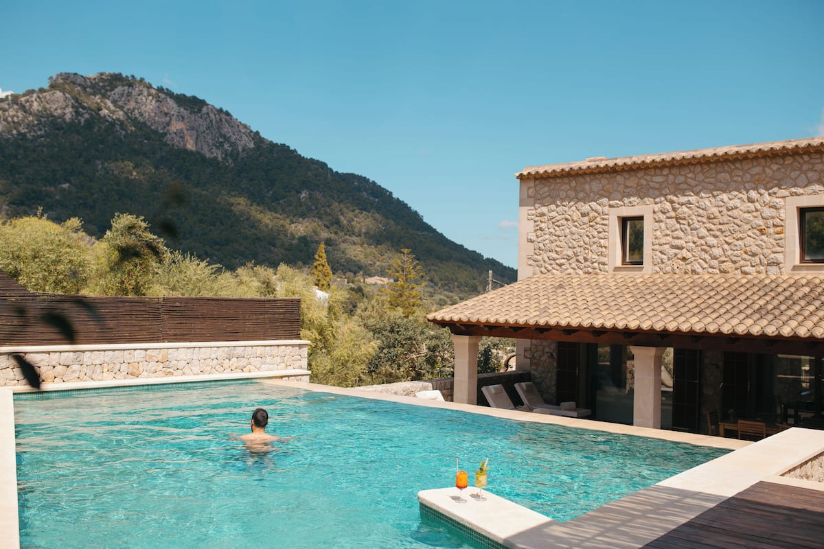 A swimming pool with a mountain backdrop, stone building, and lounge area.