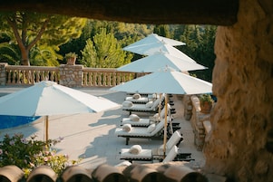 Zona junto a la piscina con sillones de descanso y sombrillas, rodeada de muros de piedra y vegetación.