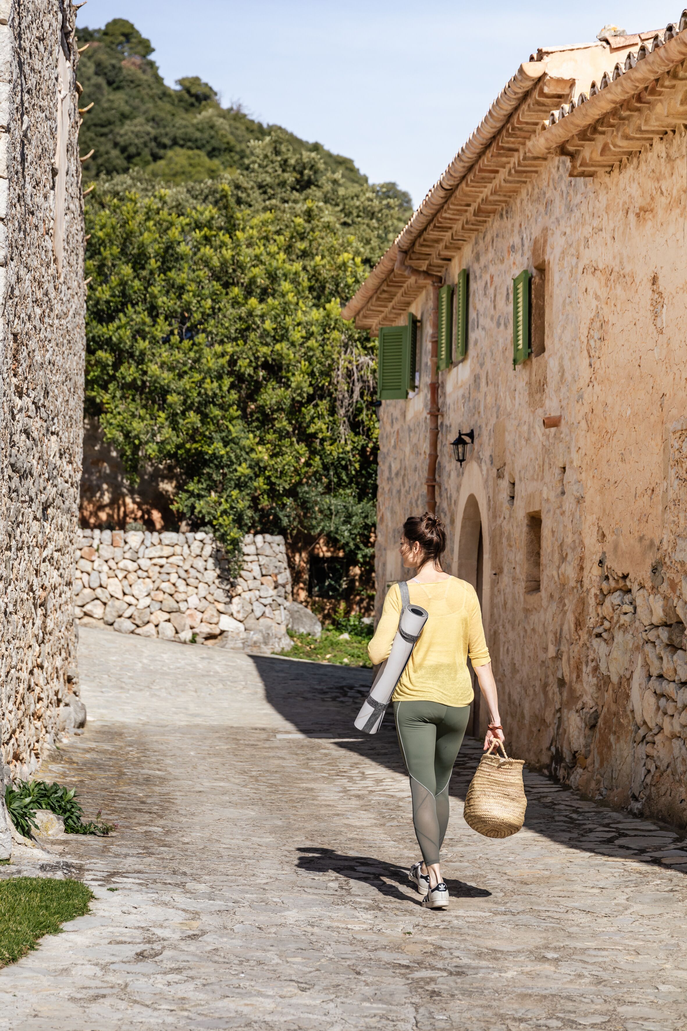 Una persona camina por un sendero empedrado con una esterilla de yoga y una cesta, pasando junto a un muro de piedra y un edificio con contraventanas verdes.