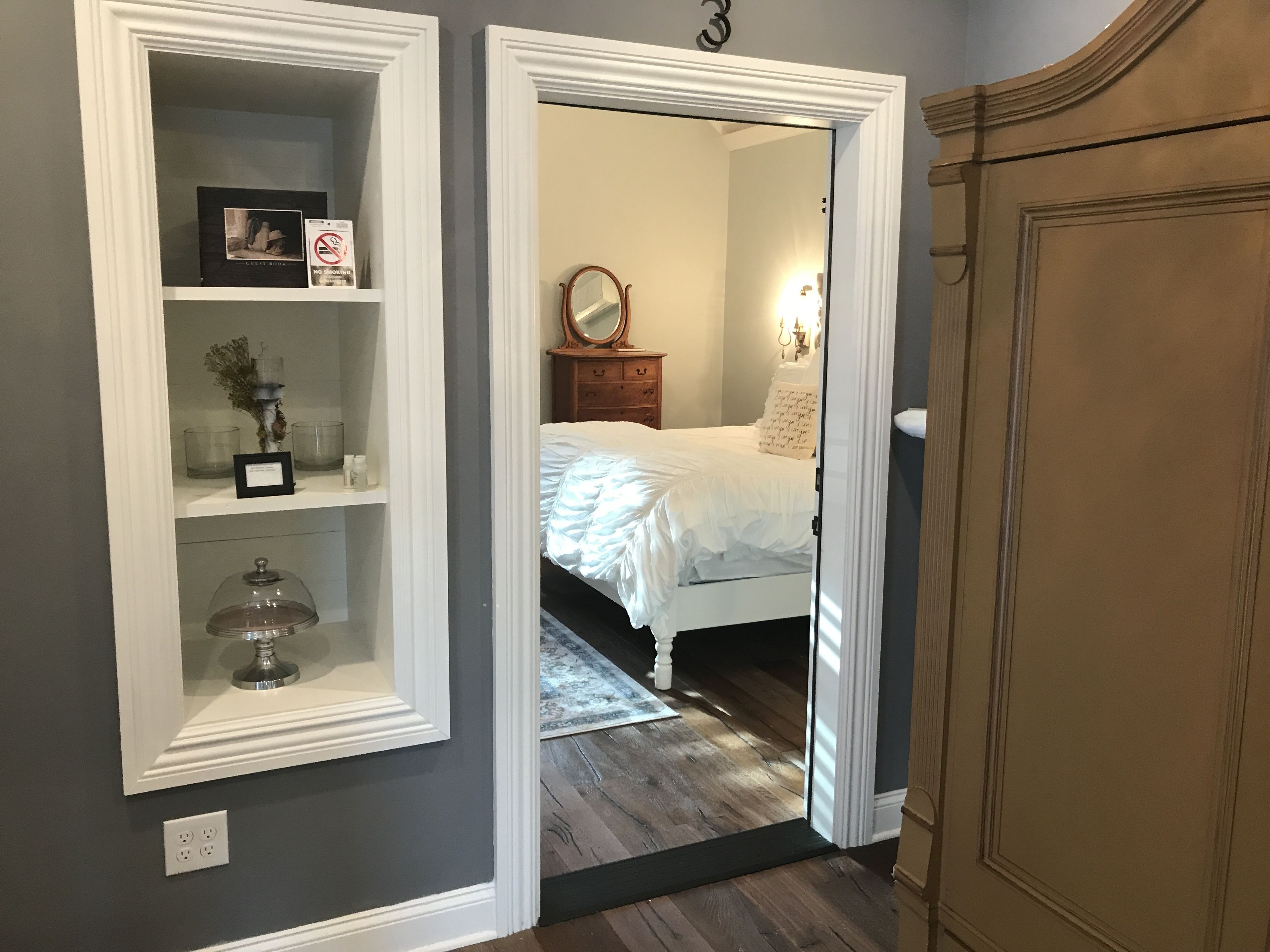 A bedroom with a bed, a dresser, and a mirror. There is a shelf with decorative items and a framed picture.