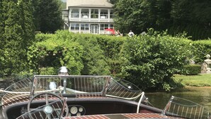 A boat docked near a house with a view of lush greenery and a building with a balcony. - Chateau Rooms (Bolton Landing)