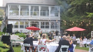 An outdoor dining area with tables and chairs, a building with large windows, and string lights in the background. - Chateau Rooms (Bolton Landing)