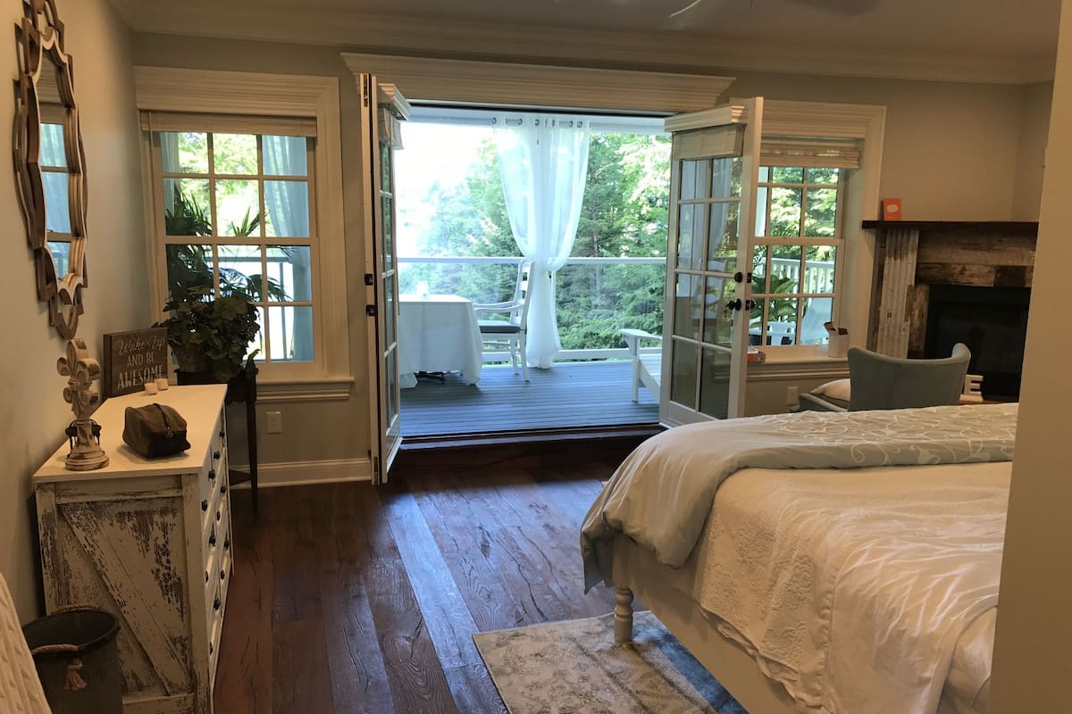 A bedroom with a large bed, a wooden floor, a view of a patio with outdoor furniture, and a fireplace.