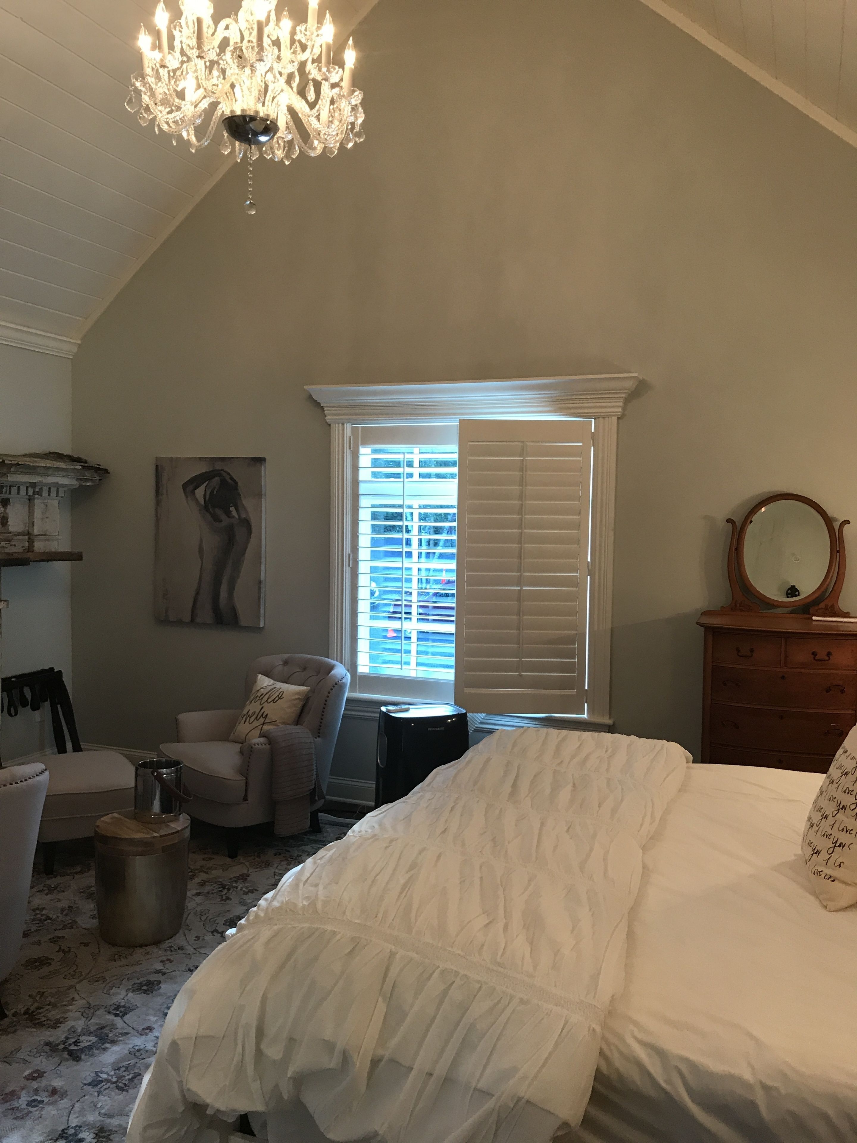 A bedroom with a large bed, a chandelier, a window with blinds, a chair, a dresser, and a painting on the wall.
