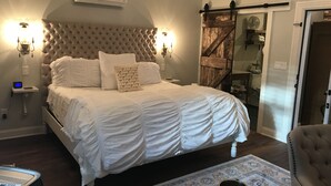 A bedroom with a tufted headboard, white bedding, a bedside table with a lamp, and a wooden door. - Chateau Rooms (Bolton Landing)