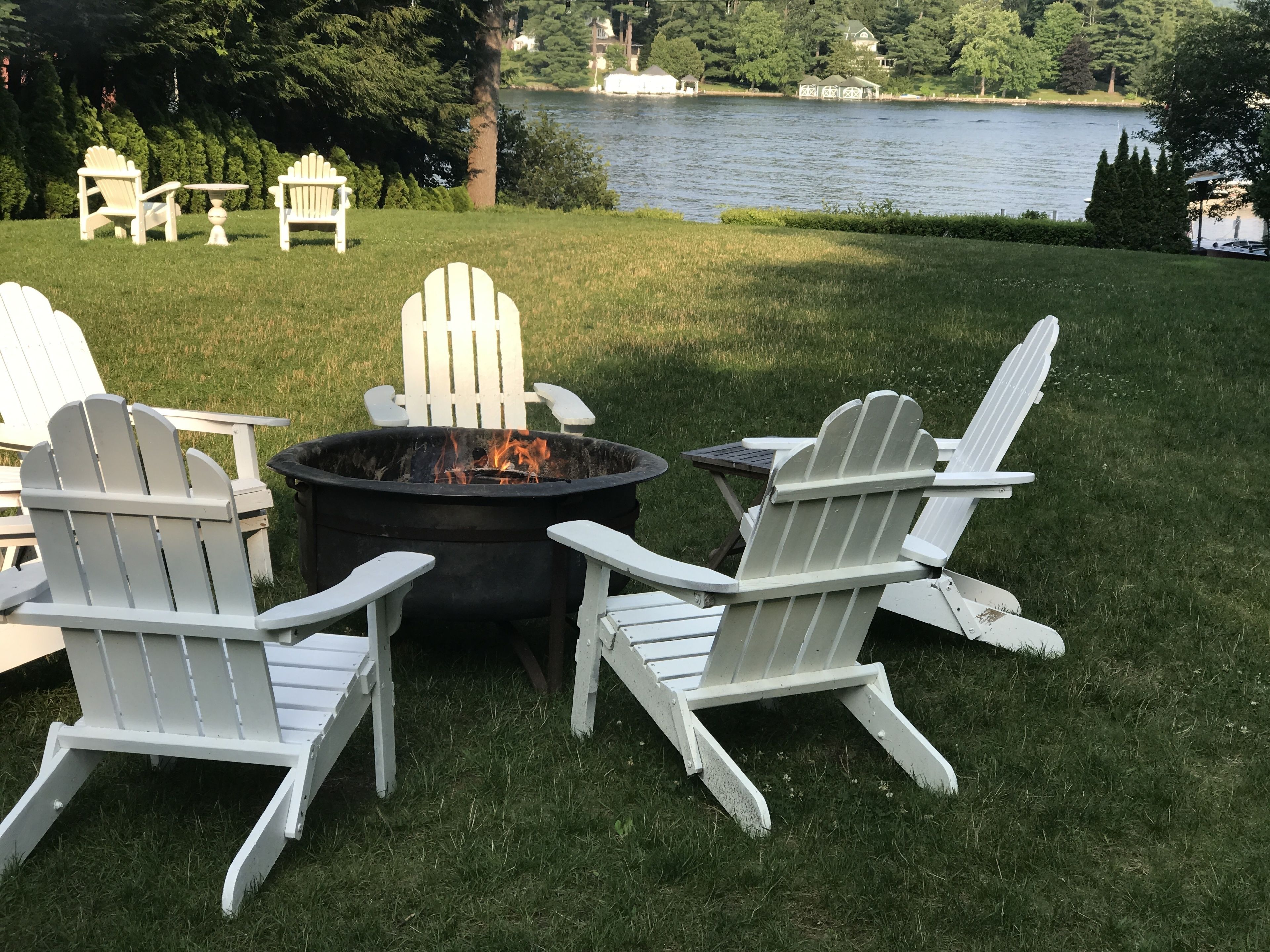 Un foyer extérieur entouré de chaises Adirondack, installé sur une pelouse verdoyante avec vue sur un lac.