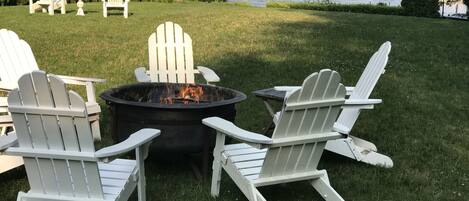 A fire pit with Adirondack chairs around it, set on a grassy lawn with a lake view.