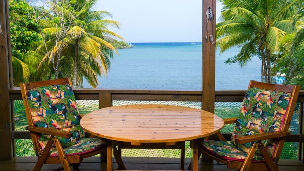 Eine Holzterrasse mit rundem Tisch und zwei Stühlen mit farbenfrohen tropischen Motiven, mit Blick auf ein ruhiges Meer und Palmen.