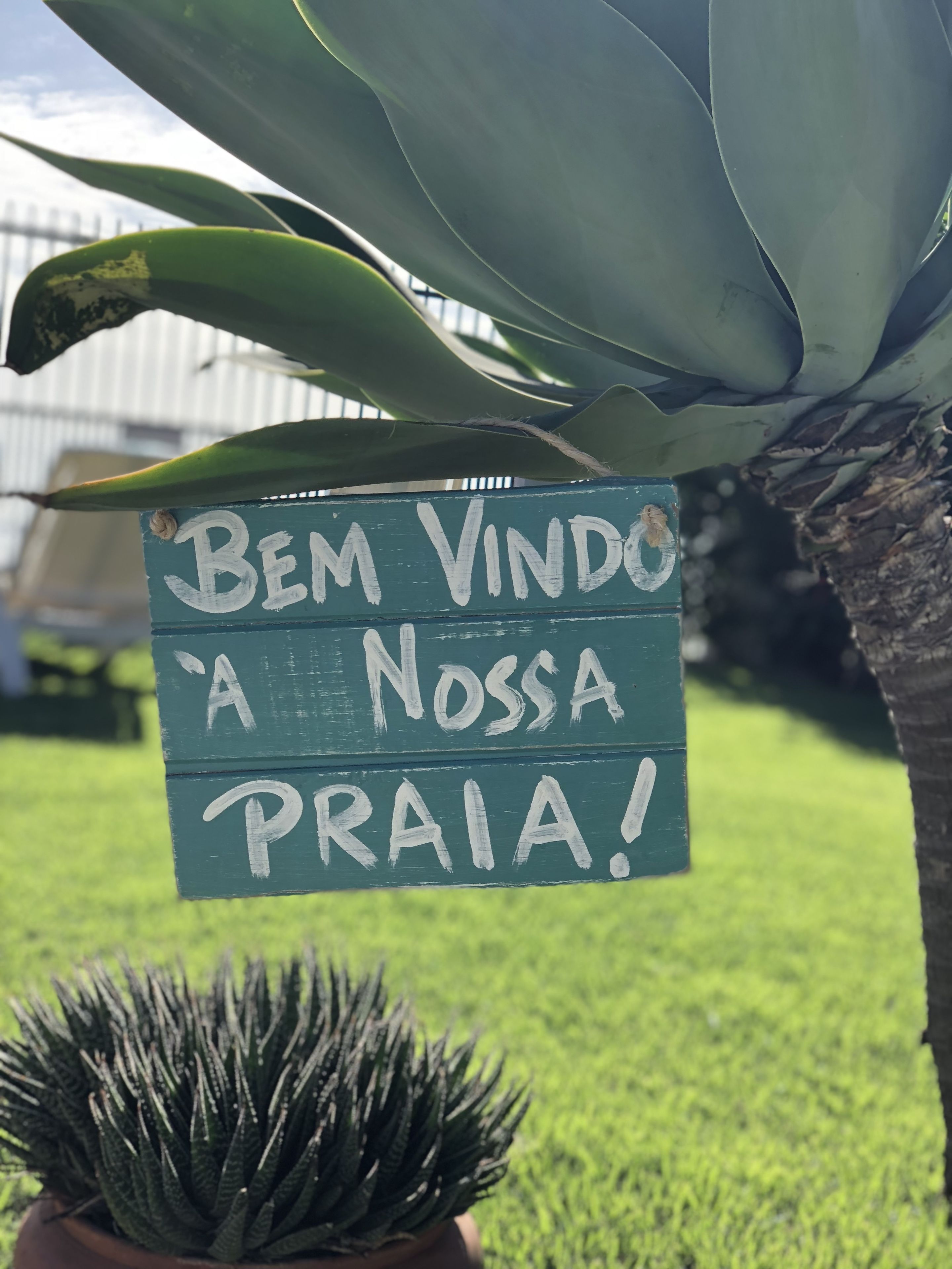 Placa com a frase 'Bem Vindo a Nossa Praia!' pendurada perto de uma planta.