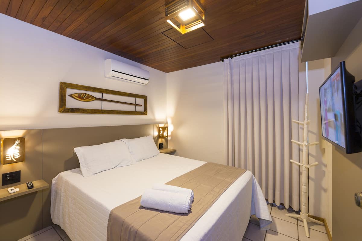 Habitación de hotel con cama, lámparas de noche, aire acondicionado, ventana con persianas y televisor montado en la pared.