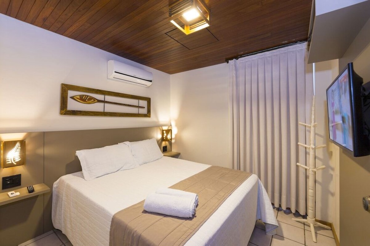 Une chambre d’hôtel équipée d’un lit, de lampes de chevet, d’un appareil de climatisation, d’une fenêtre avec stores et d’un téléviseur fixé au mur.