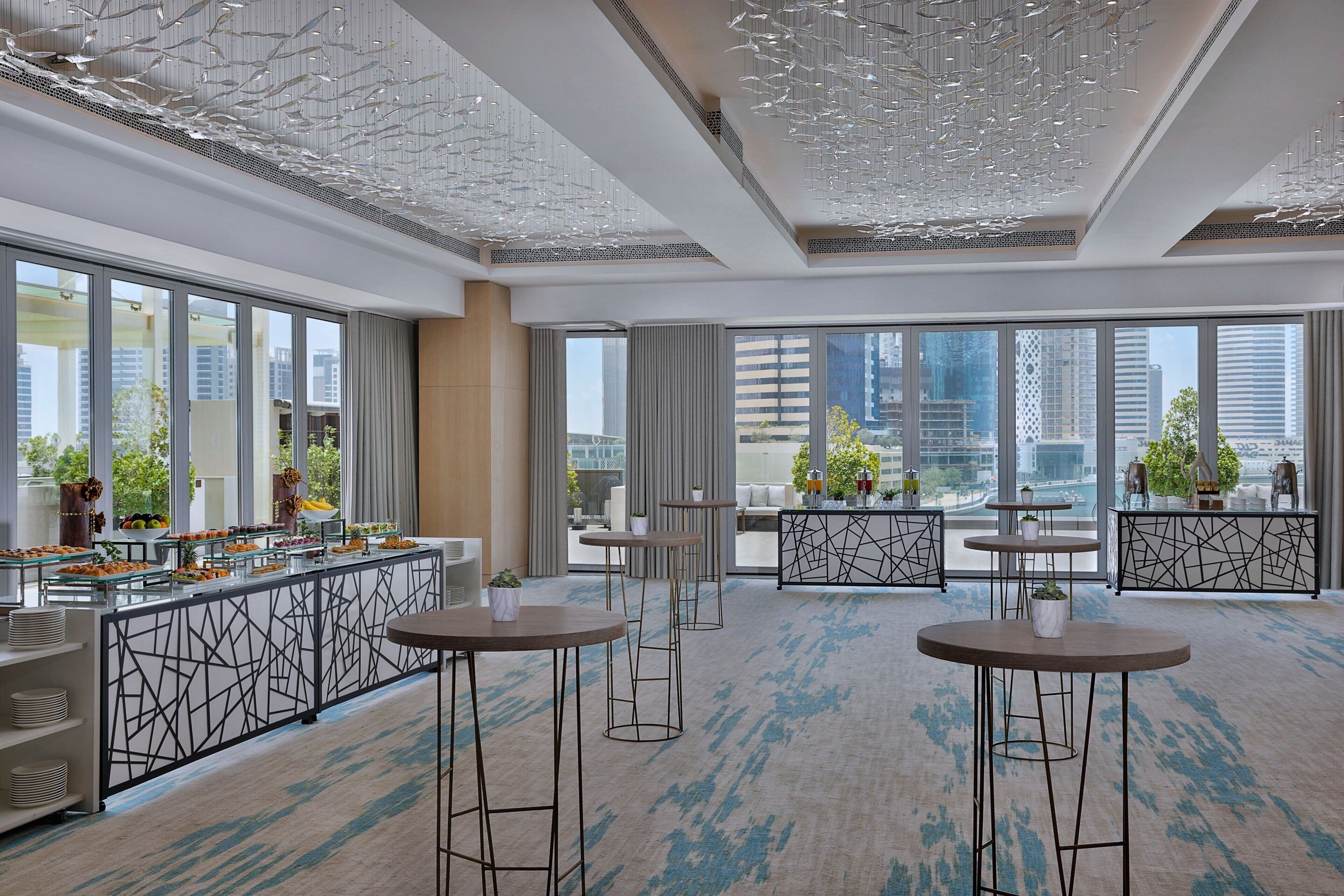 A modern hotel lobby with a high ceiling, large windows offering city views, a buffet area, and several round tables with stools.