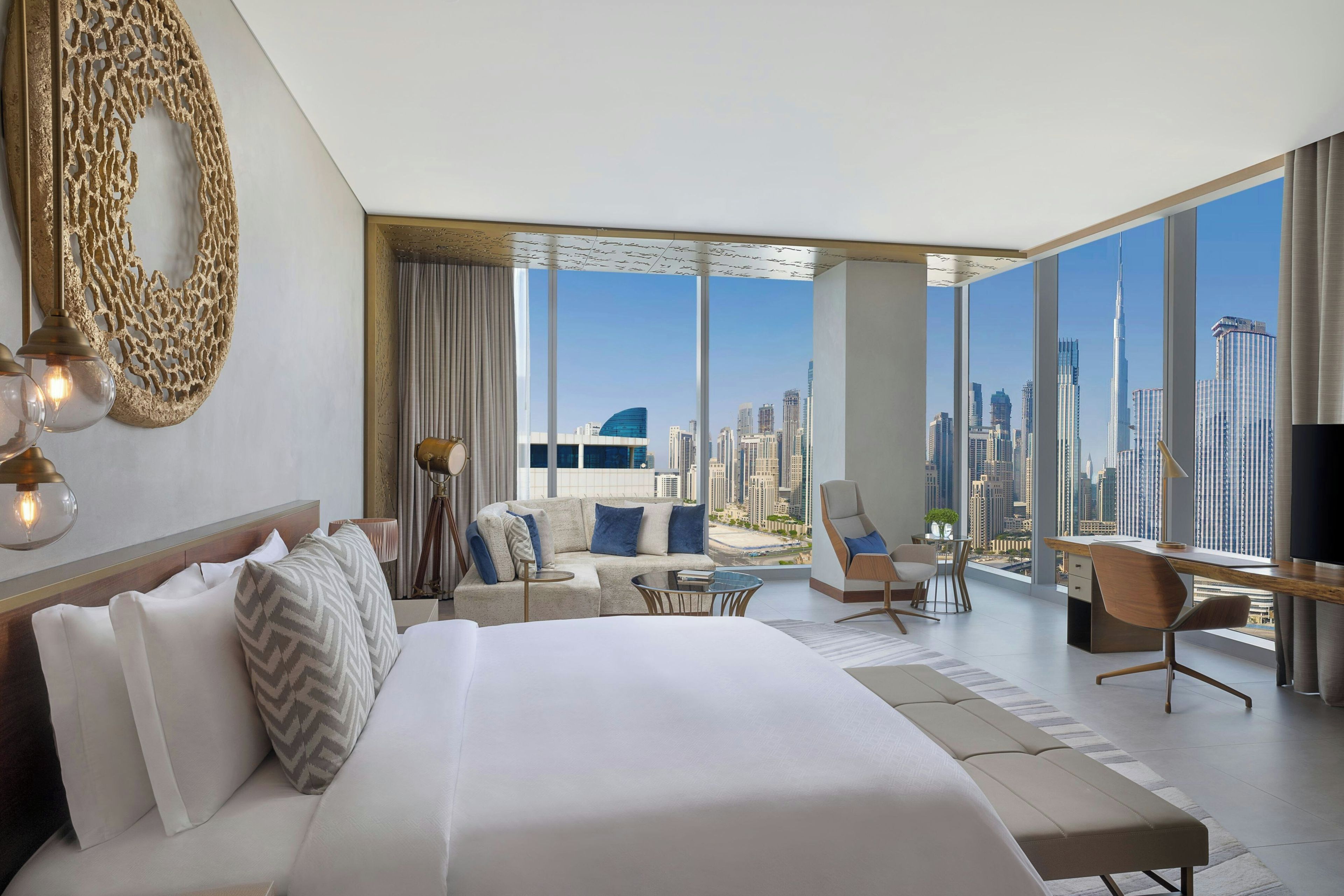 A modern hotel room with a large bed, a desk, a chair, and a view of a cityscape.
