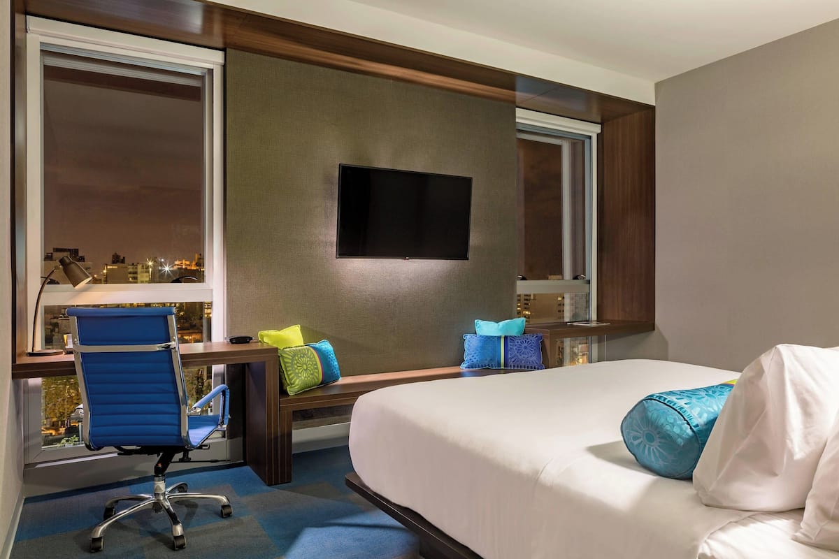 A hotel room with a bed, a desk with a chair, a flat-screen TV, and a window with a city view.