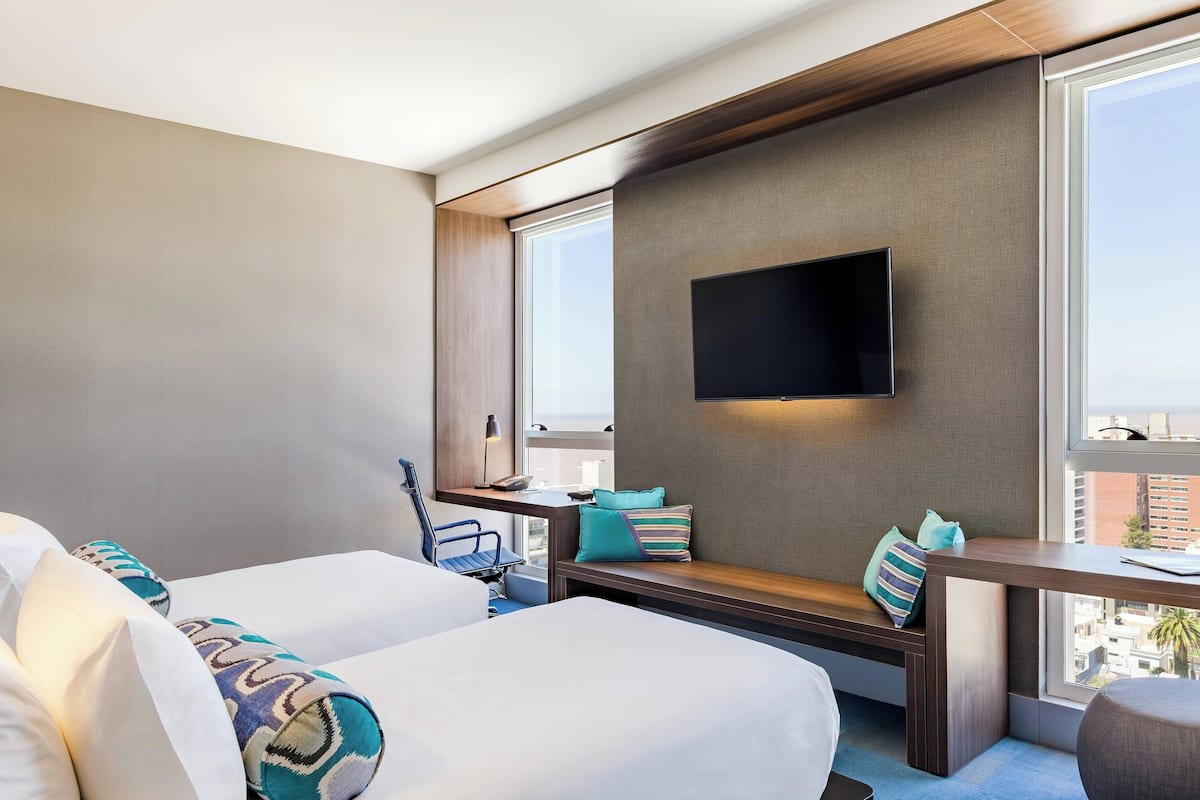 A hotel room with two beds, a TV mounted on the wall, a desk with a chair, and a window with a city view.