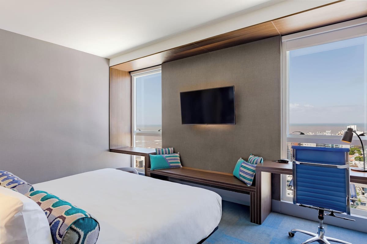 A modern hotel room with a large bed, a desk with a chair, a flat-screen TV mounted on the wall, and a view of the city through large windows.