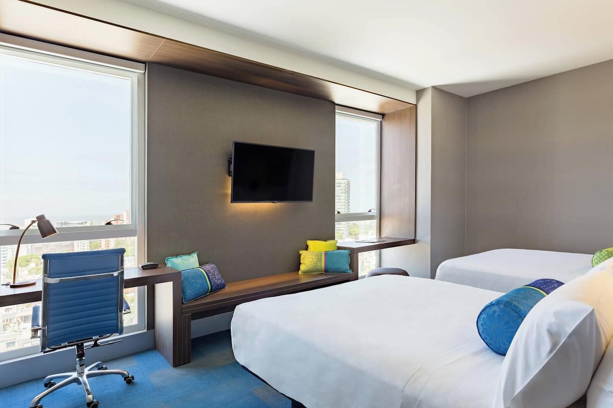 A hotel room with a large window, a desk with a flat-screen TV, two beds, and a chair.