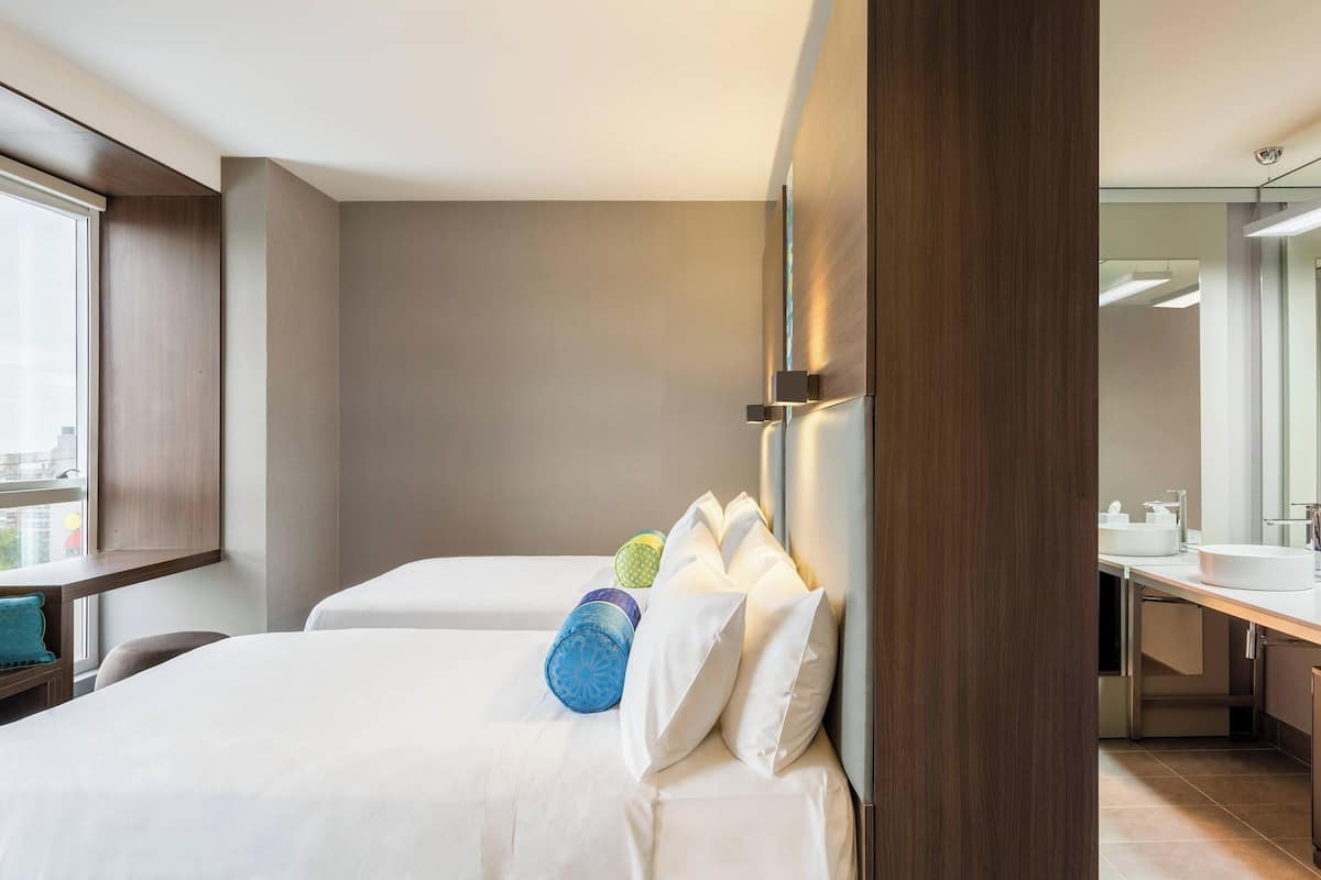 A hotel room with two beds, a large window, a modern bathroom, and a view of the city.