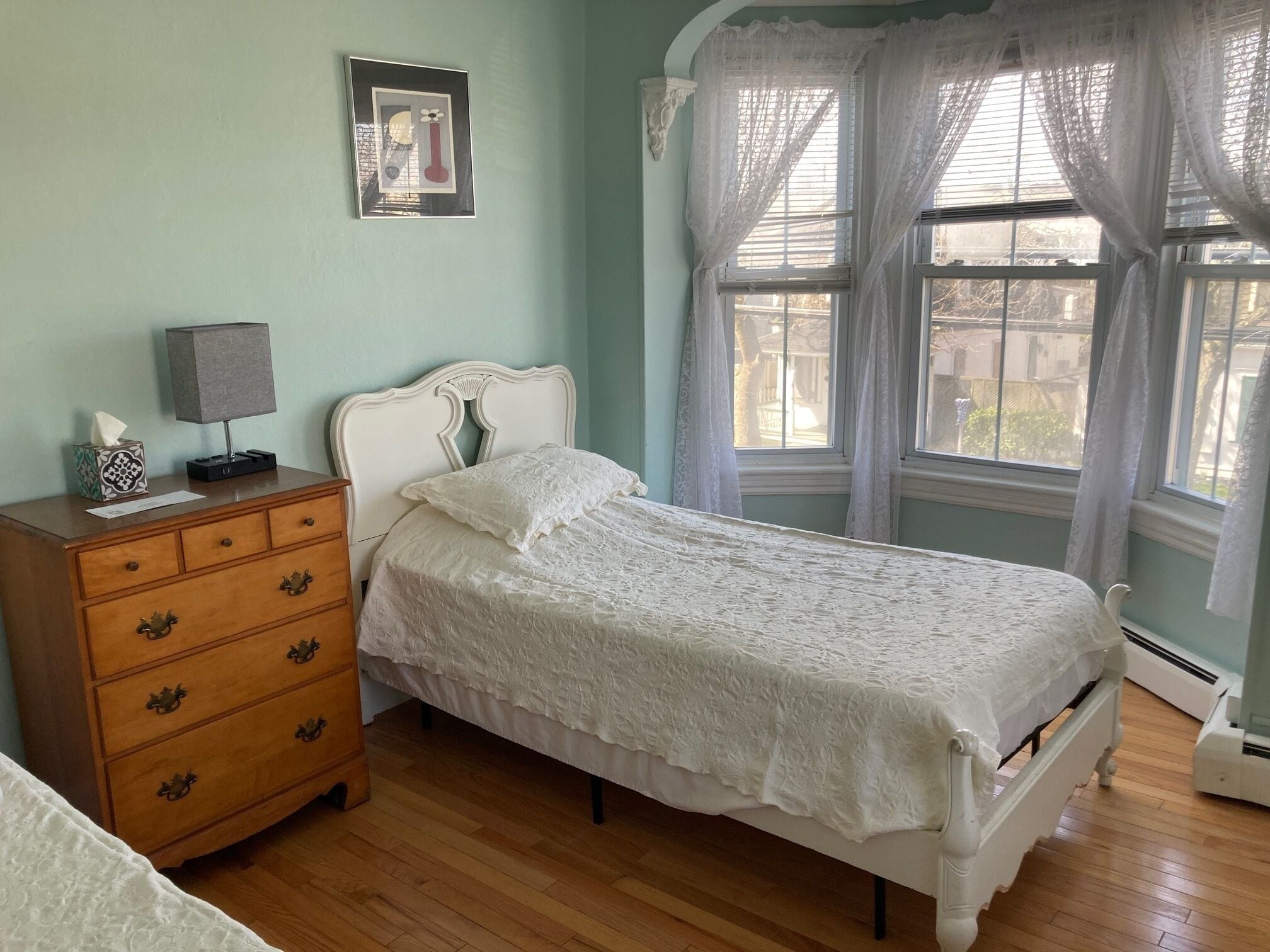 A bedroom with a wooden dresser, a white bed with a lace cover, a framed picture on the wall, and a window with sheer curtains.