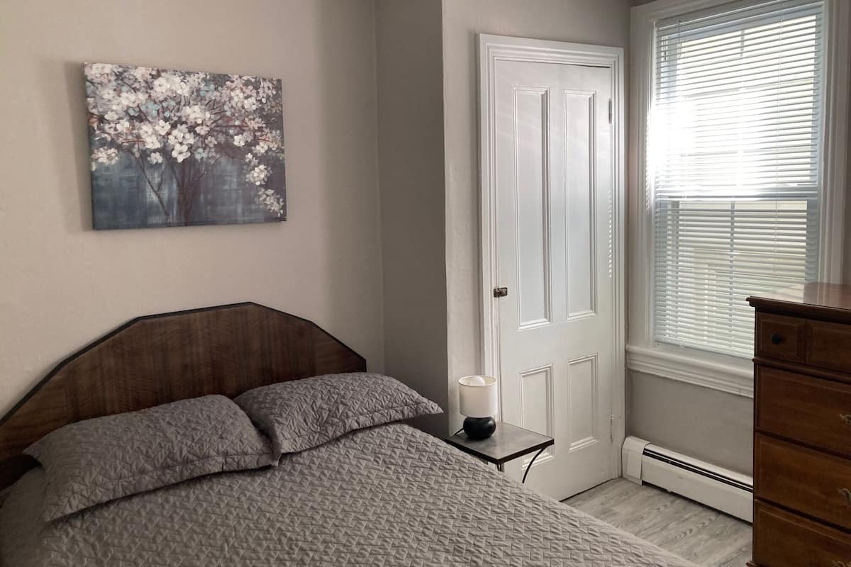 A bedroom with a bed, a dresser, a door, and a window with blinds.