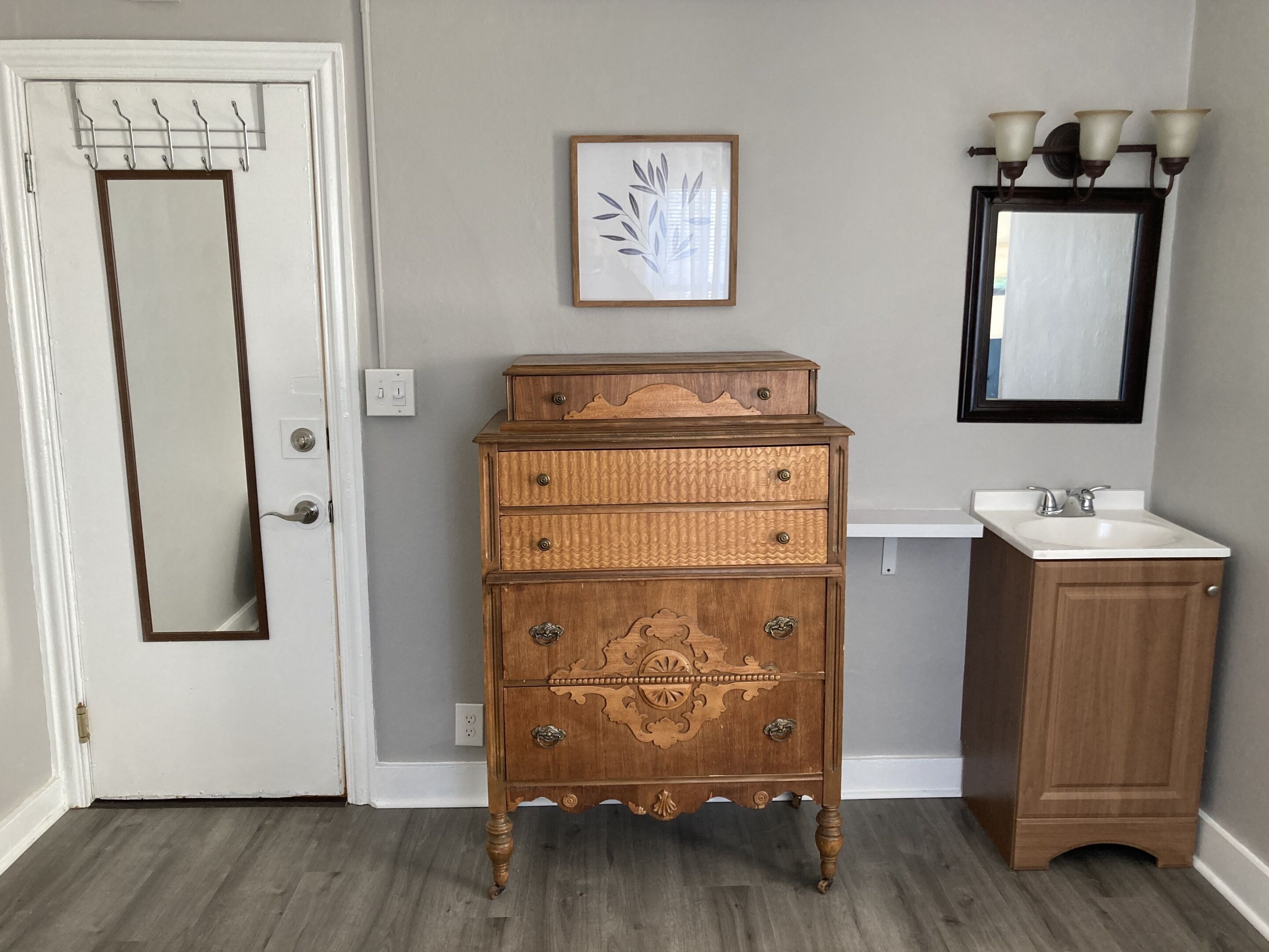 A wooden dresser with drawers and a mirror above it, a small sink, and a door with a coat rack.