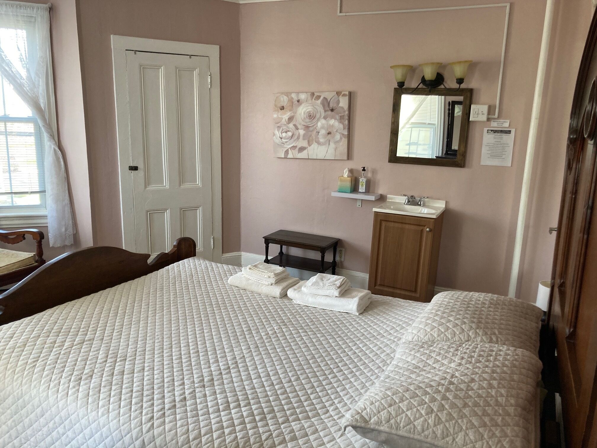 Un dormitorio con una cama grande, una silla de madera, una mesita, un espejo y un estante en la pared con artículos de aseo.