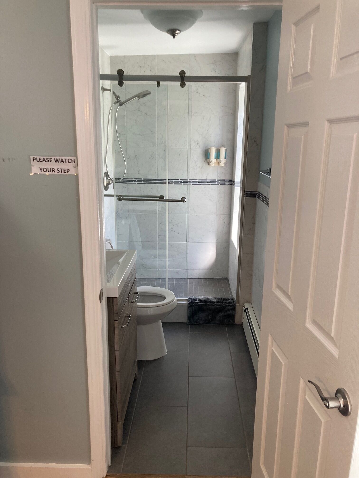 A bathroom with a toilet, shower, and a sign that says "PLEASE WATCH YOUR STEP".