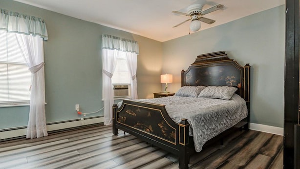 A bedroom with a dark wooden bed, a ceiling fan, and a window with white curtains. - Ocean Park Inn (Ocean Grove)