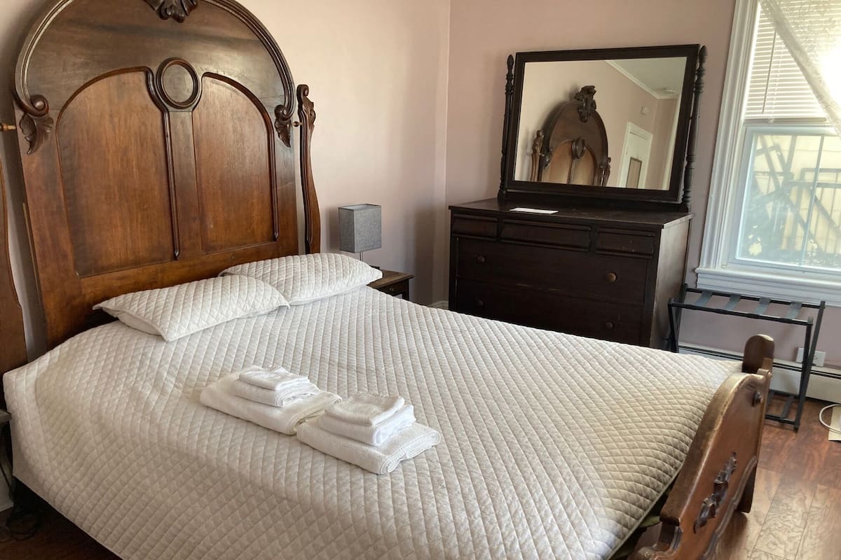 A neatly made bed with a quilted white bedspread and pillows, a wooden headboard, a dark dresser with a mirror, and a window with blinds.