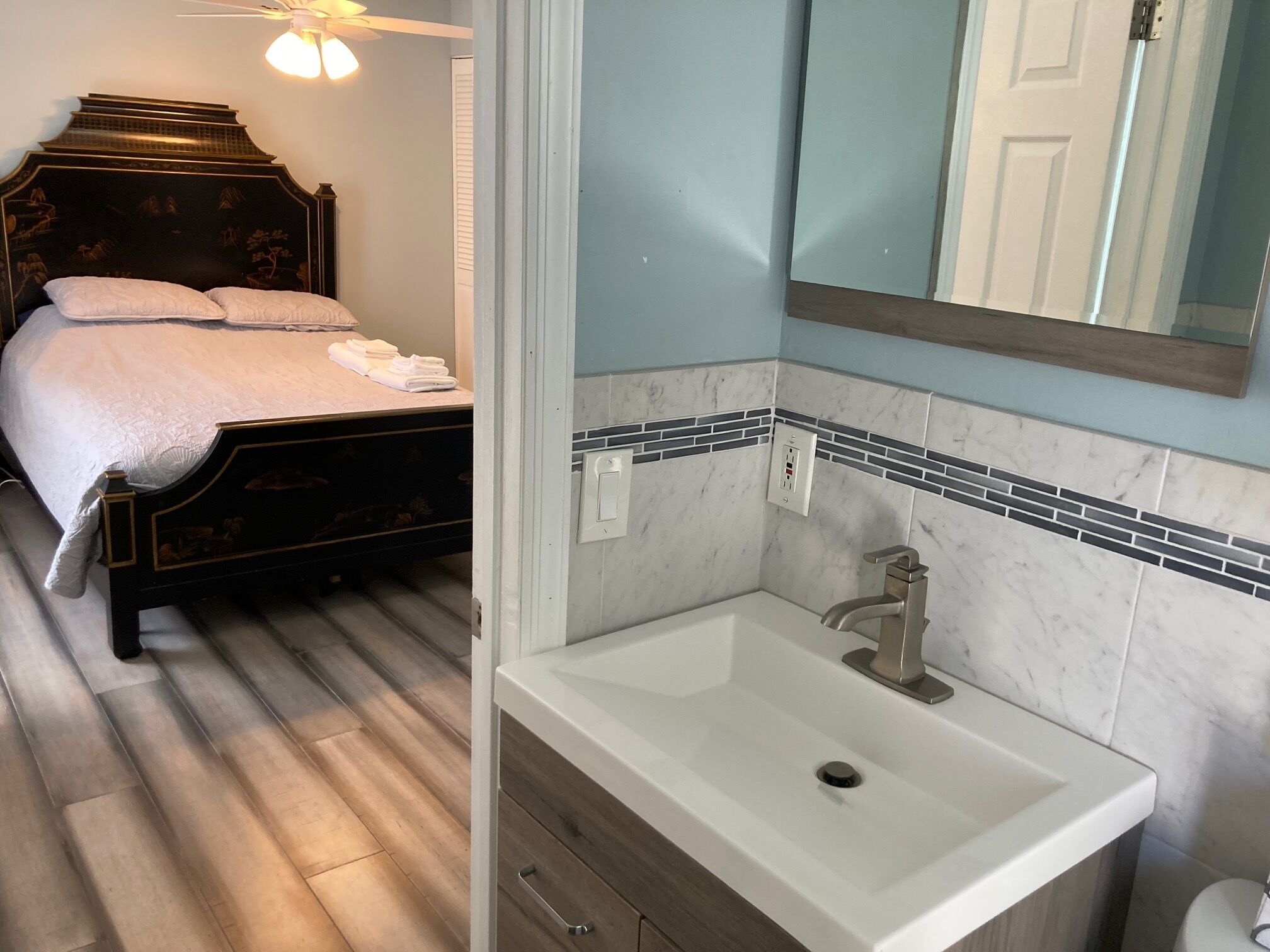 Un dormitorio con cama, mesita de noche, ventilador de techo y baño con lavamanos, espejo y salpicadero de azulejos.