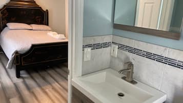 Un dormitorio con cama, mesita de noche, ventilador de techo y baño con lavamanos, espejo y salpicadero de azulejos.
