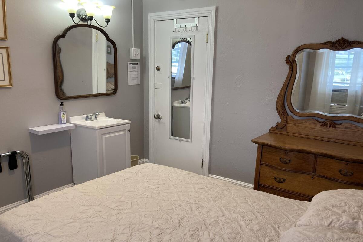 A bedroom with a bed, a dresser, a vanity with a mirror, and a door with a coat rack.