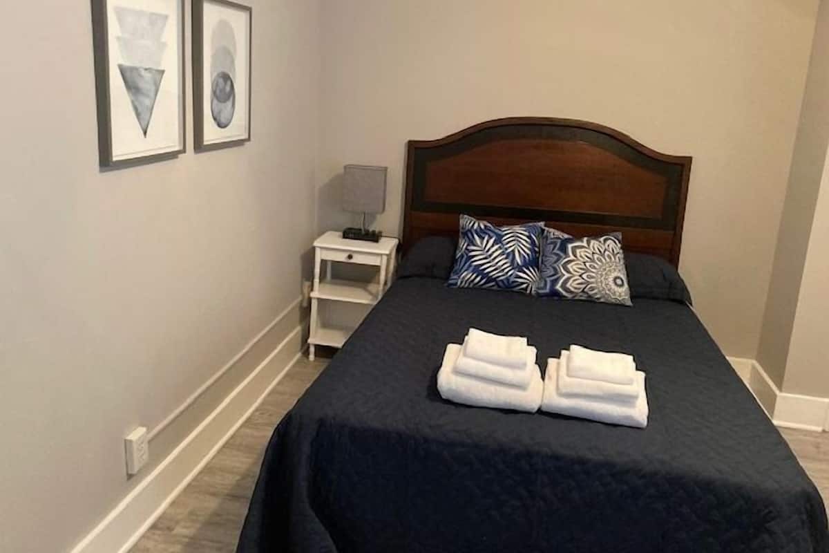 A neatly made bed with a dark blue bedspread and two pillows, a small bedside table with a lamp, and two framed pictures on the wall.