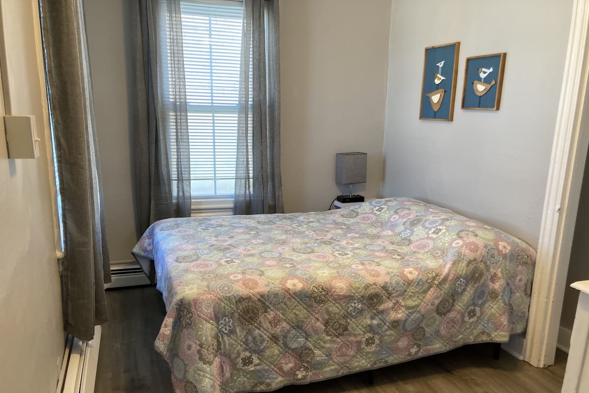 A bedroom with a floral-patterned bedspread, a window with sheer curtains, and two wall-mounted bird-themed decorations.