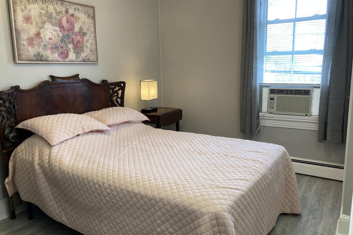 A neatly made bed with a quilted bedspread, a wooden headboard, a bedside table with a lamp, and a window with curtains.